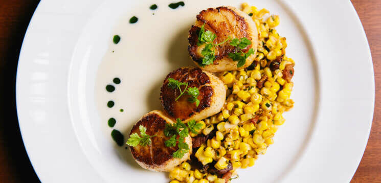 A white plate with 3 scallops and corn on a plate.