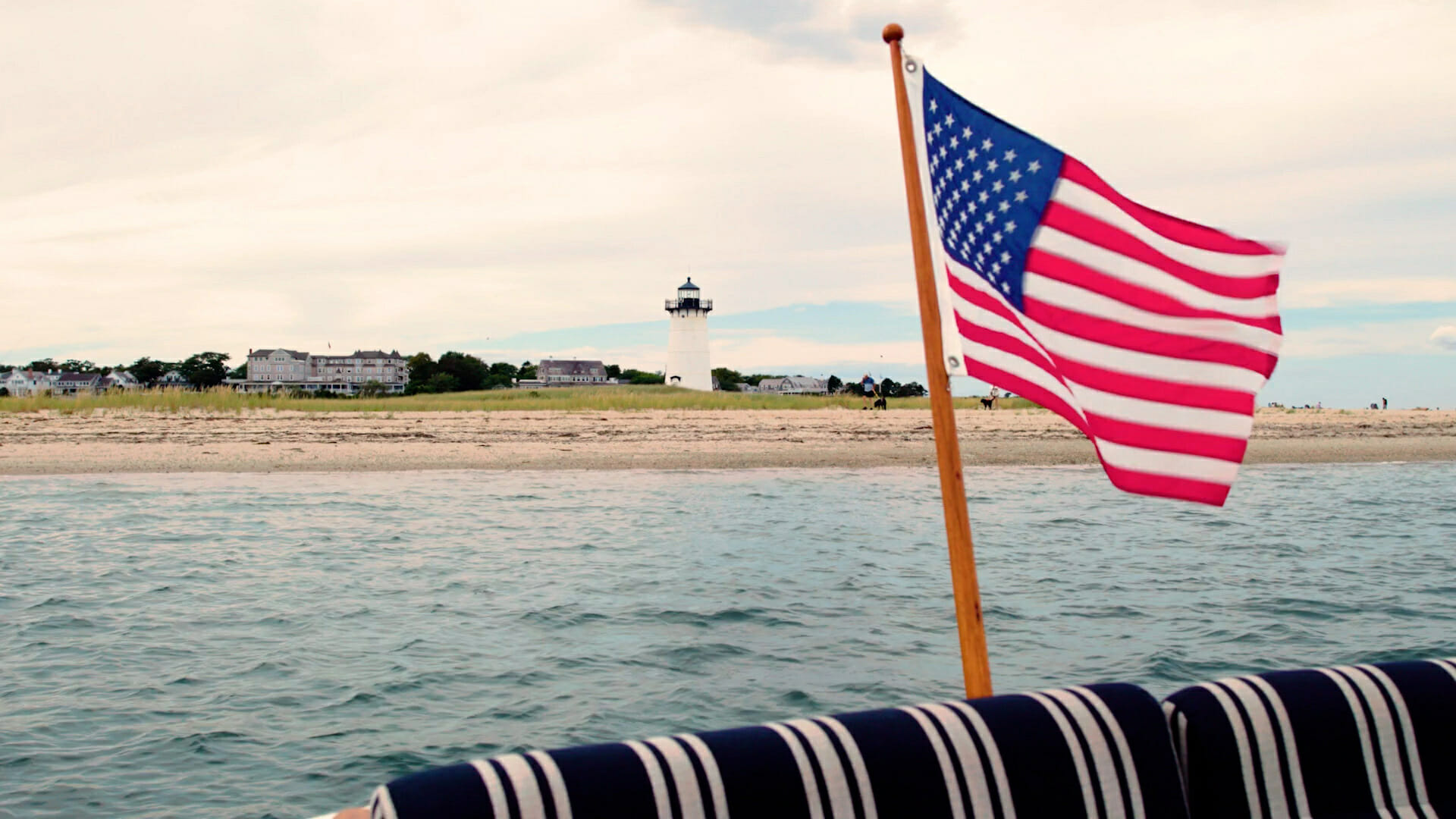 American flag in the foreground of blue-grey water surrounding an island with a lighthouse