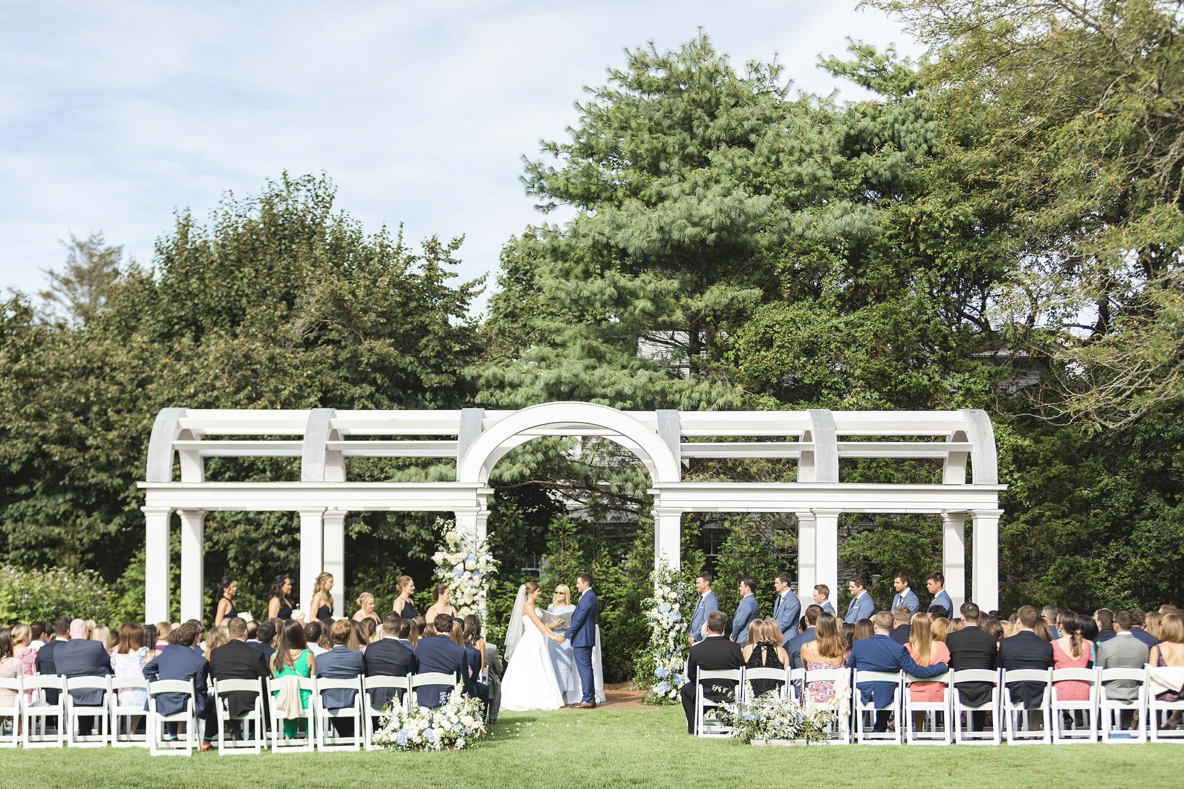 white chairs and people sitting in them with a couple standing in front of a white pergola