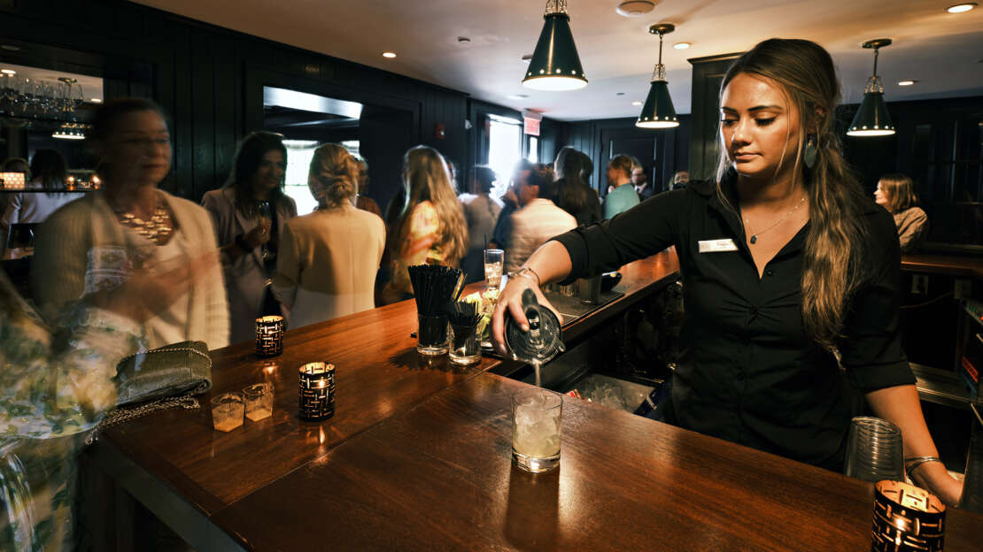 a woman bartender pouring a drink at the bar and a group of people standing and talking