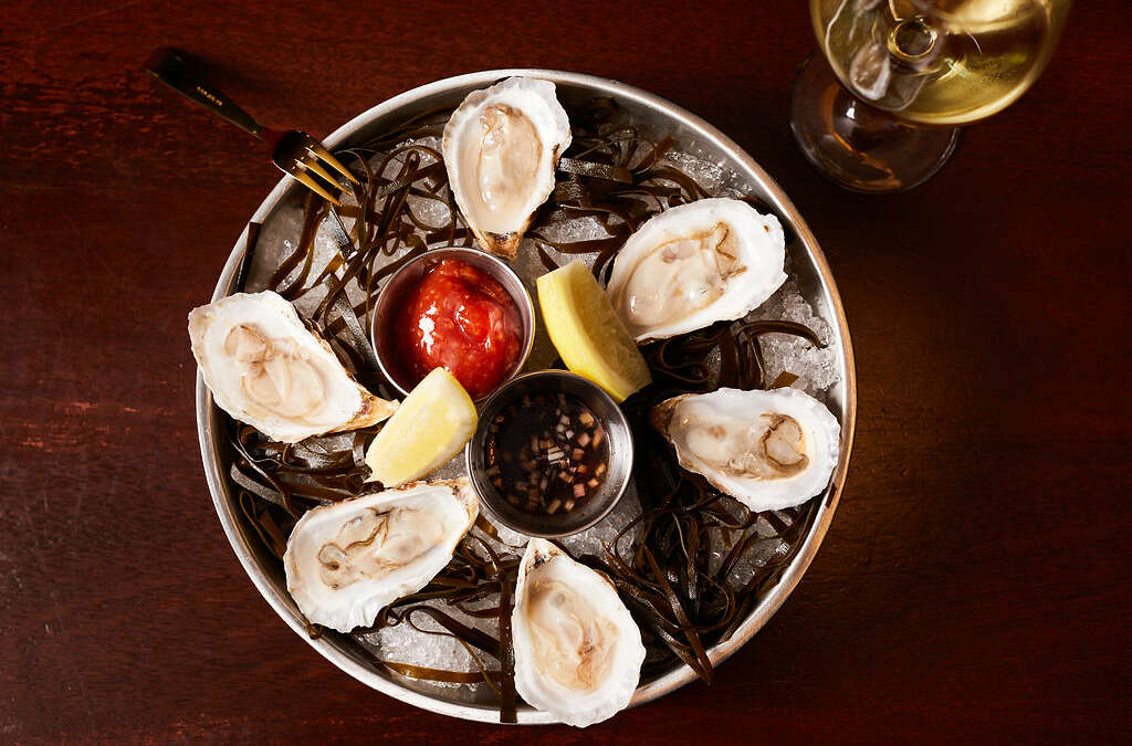 six oysters in a silver bowl with ice and seaweed and a gold fork