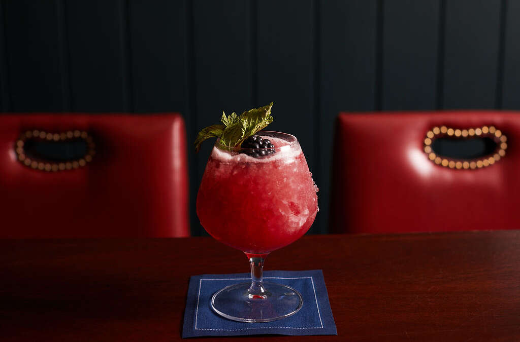 a red drink with a mint garnish, blackberry, on a blue napkin and two red chairs
