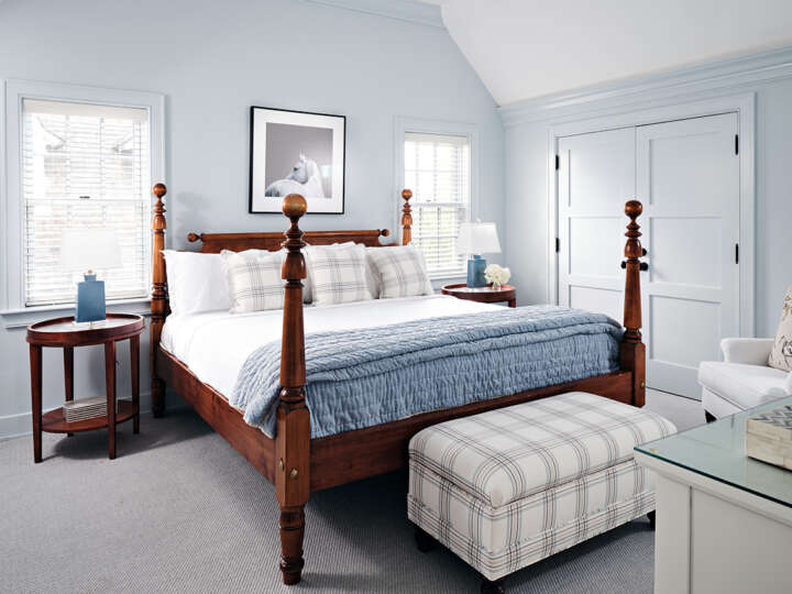 a bedroom with a four-poster bed, with a blue and white blanket and a cream-colored bench at the end of the bed