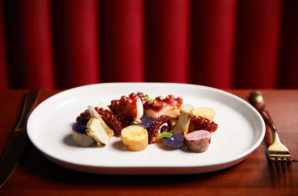 a white plate with cooked octopus, potatoes, with a red background and fork and knife