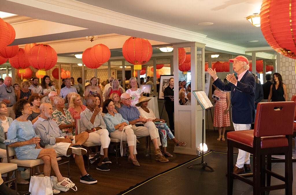 A large crowd of people sitting with red chinese lanterns hanging from the ceiling. A man talking with a red hat.